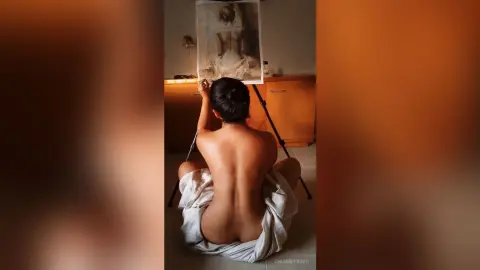 Nude woman sitting in a dimly lit room looking at a portrait painting on an easel.