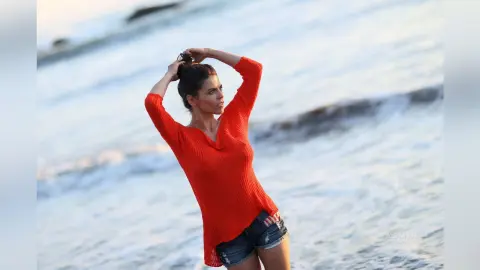 Beautiful woman in a bright orange sweater and denim shorts standing on a breezy beach at sunset.