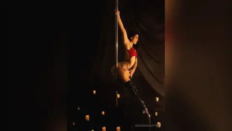 Athletic woman in red bodysuit and black boots performing pole dance in a moody, dark studio setting.
