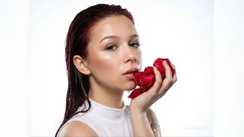 Young woman with wet red hair holding a red rose against a white background in a soft, romantic mood.