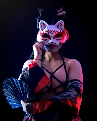 Woman in red and black kimono wearing a white kitsune mask and holding a blue fan in moody lighting.
