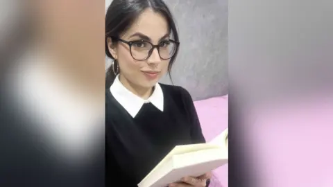 Young woman with glasses and black sweater reading a book in a cozy indoor setting.