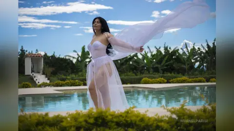 Dreamy woman in a flowing white sheer gown standing by a luxury pool under a blue sky.