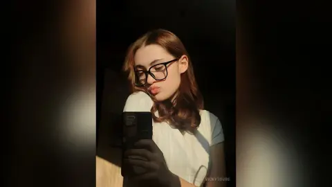 VickyToure wearing black glasses and a white t-shirt, taking a mirror selfie with her phone in dim lighting.