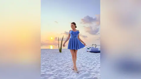 Young woman in a blue summer dress walking on a white sand beach during a dreamy tropical sunset.