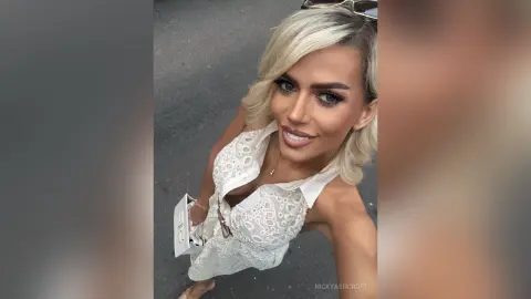 Smiling blonde woman in a white lace sleeveless blouse posing outdoors in bright daylight.