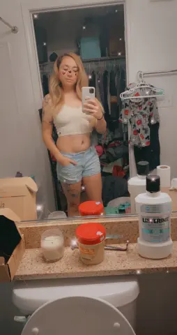 Young woman in a white crop top and denim shorts taking a mirror selfie in a bright bathroom.