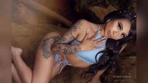 Tattooed woman in a light blue bikini relaxing on rocks in a moody, natural outdoor setting.