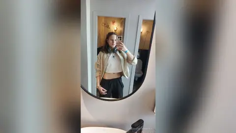 Young woman in a beige crop top and black joggers taking a mirror selfie in a modern bathroom.