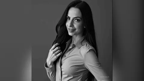 Smiling woman with long dark hair wearing a light button-down shirt in a moody black and white studio portrait.