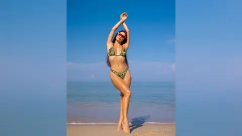 Fashionable woman in a green patterned bikini posing on a sunny tropical beach under a clear blue sky.