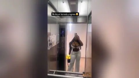 Stylish woman in a white crop top and light trousers taking a mirror selfie in an elevator.
