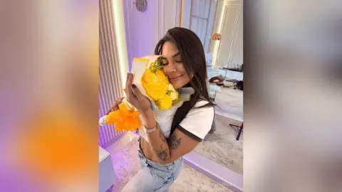 Smiling young woman in a white crop top hugging a large bouquet of yellow roses in a bright modern room.