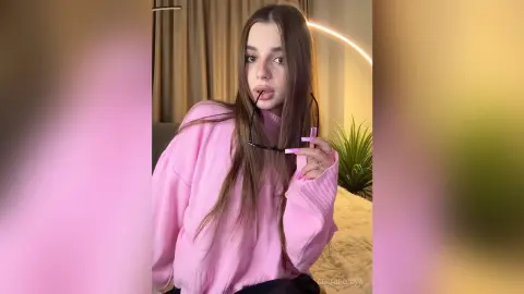 GertudeCova wearing a pink sweatshirt, posing with her glasses down and long brown hair visible in a room with a light ring and plant.