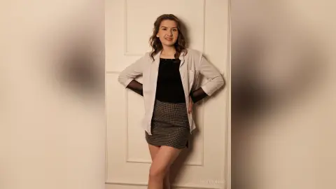 Smiling young woman in a white shirt and black top posing against a white door in a bright indoor setting.