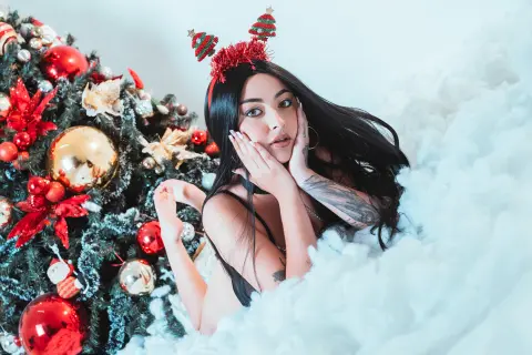 Glamorous woman in festive reindeer ears relaxing by a decorated Christmas tree in a dreamy snowy setting.