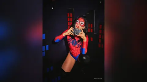 JessieLiu poses in a red and blue Spider-Man bodysuit while holding a black camera in a dark room.