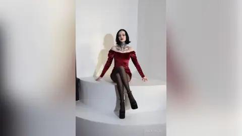 Gothic woman in a red velvet dress and black tights sitting in a minimalist white studio setting.