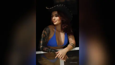 Tattooed woman in a blue bikini and black cowboy hat posing outdoors in soft natural light.