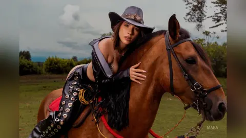 Fashionable woman in black leather outfit riding a brown horse in a scenic outdoor field at sunset.