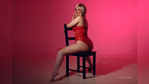 KaseyLang poses sitting sideways on a black chair wearing a red lace bodysuit and white patterned thigh-high stockings.