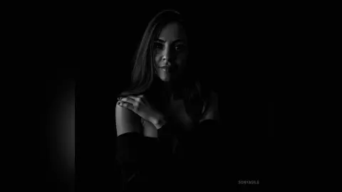 Moody black and white portrait of a woman in a dark outfit against a shadowy background.