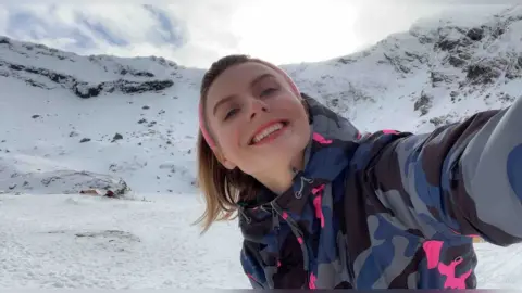 Smiling woman in a pink headband and camo winter jacket enjoying a sunny day in the snowy mountains.