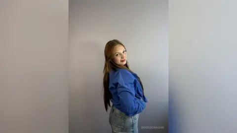 DorisDonachie poses in a blue long-sleeve shirt and denim shorts against a plain grey wall.
