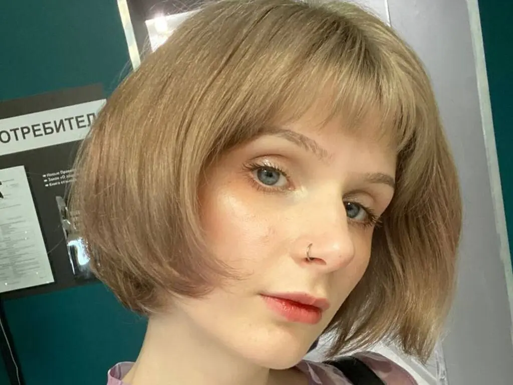 AlitaPatti looks at the camera with a short blonde bob haircut and a nose ring, wearing a light pink top.