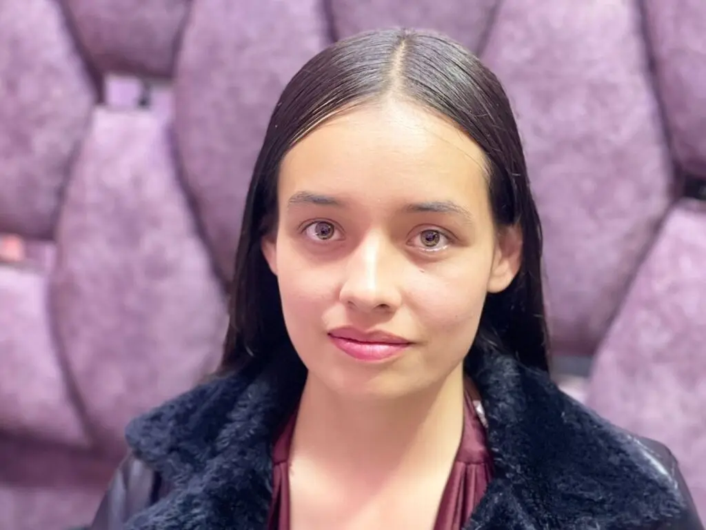 Young woman with long dark hair wearing a black faux fur coat against a textured purple background.