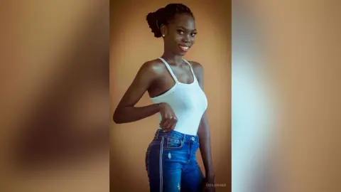 Smiling young woman in a white tank top and blue jeans posing against a neutral studio background.
