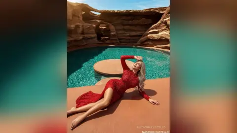 NatashaCastello poses in a tight red dress while lounging by a turquoise pool in a desert canyon setting.