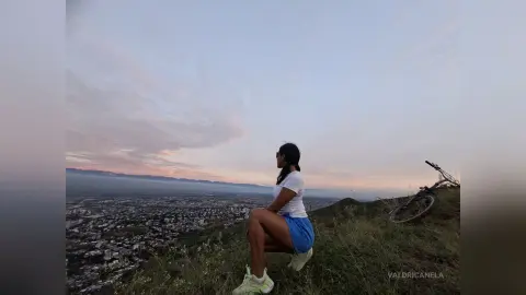 Young woman in white t-shirt and blue shorts sitting on a grassy hill overlooking a city at sunset.