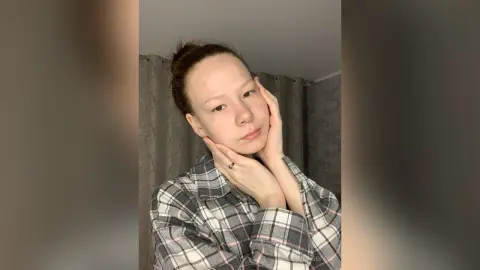 Young woman in a grey plaid shirt posing thoughtfully in a cozy indoor setting.