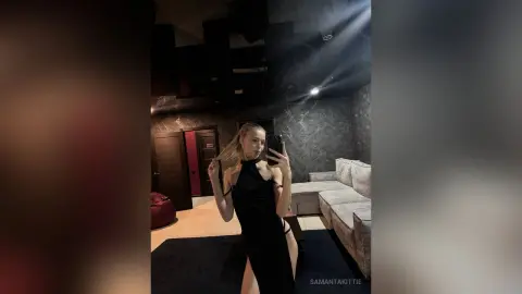 Blonde woman in a chic black halter dress taking a mirror selfie in a moody, modern dark living room.