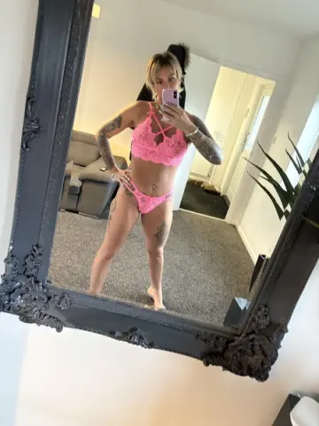 Tattooed woman in pink lace lingerie taking a mirror selfie in a bright modern bedroom.