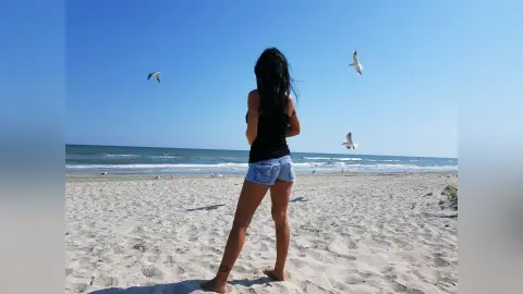 Woman in black tank top and denim shorts standing on a sunny beach looking at seagulls over the ocean.