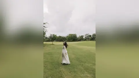 StacyHarland stands in a white long-sleeved dress on a wide green lawn under a cloudy sky.
