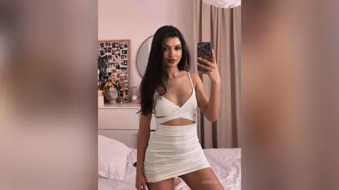 Young woman in a white cutout mini dress taking a mirror selfie in a bright bedroom.