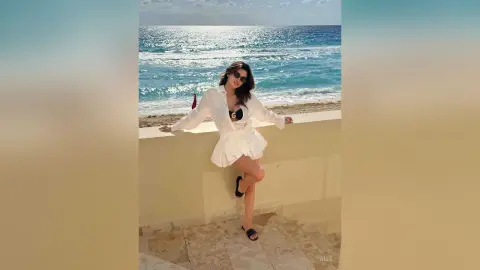 Stylish woman in a white long-sleeve romper and sunglasses relaxing by the ocean on a sunny beach day.