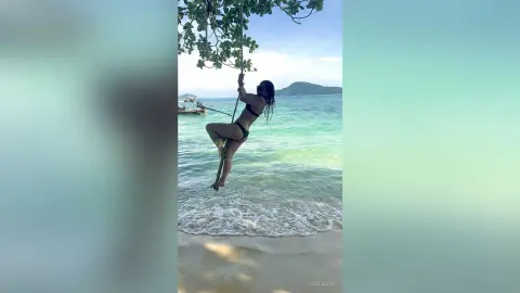 Woman in black bikini swinging on a rope over turquoise tropical ocean waves under a bright summer sky.