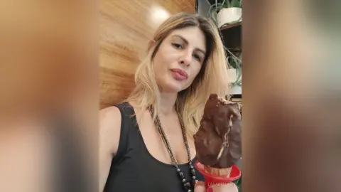 Blonde woman in a black tank top holding a chocolate-covered ice cream bar in a bright indoor setting.