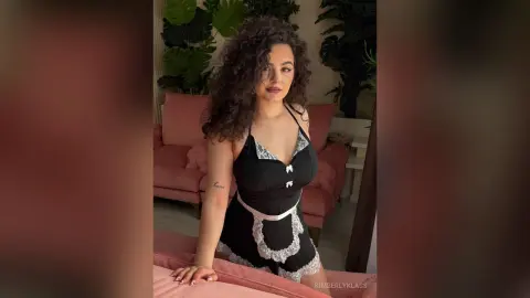 Woman with curly hair in a black lace romper sitting on a pink sofa in a bright, cozy living room.