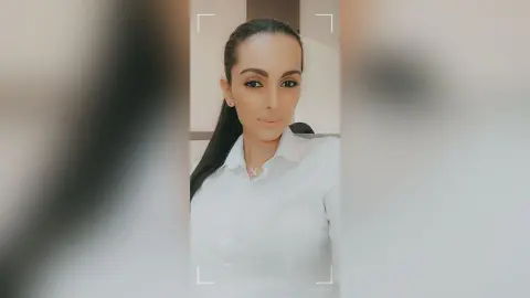 AishaApril poses in a white collared shirt with her long dark hair pulled back, looking directly at the camera.