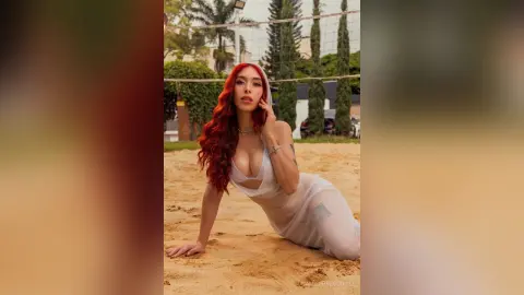 Red-haired woman in a white sheer beach cover-up posing on a sunny sand volleyball court.