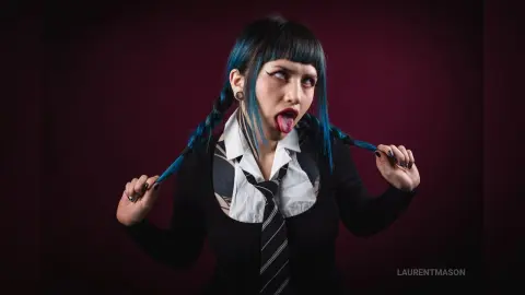 Gothic woman with blue hair and black outfit sticking her tongue out against a dark moody background.