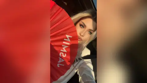 Blonde woman in a white top holding a red fan inside a car during golden hour sunset.
