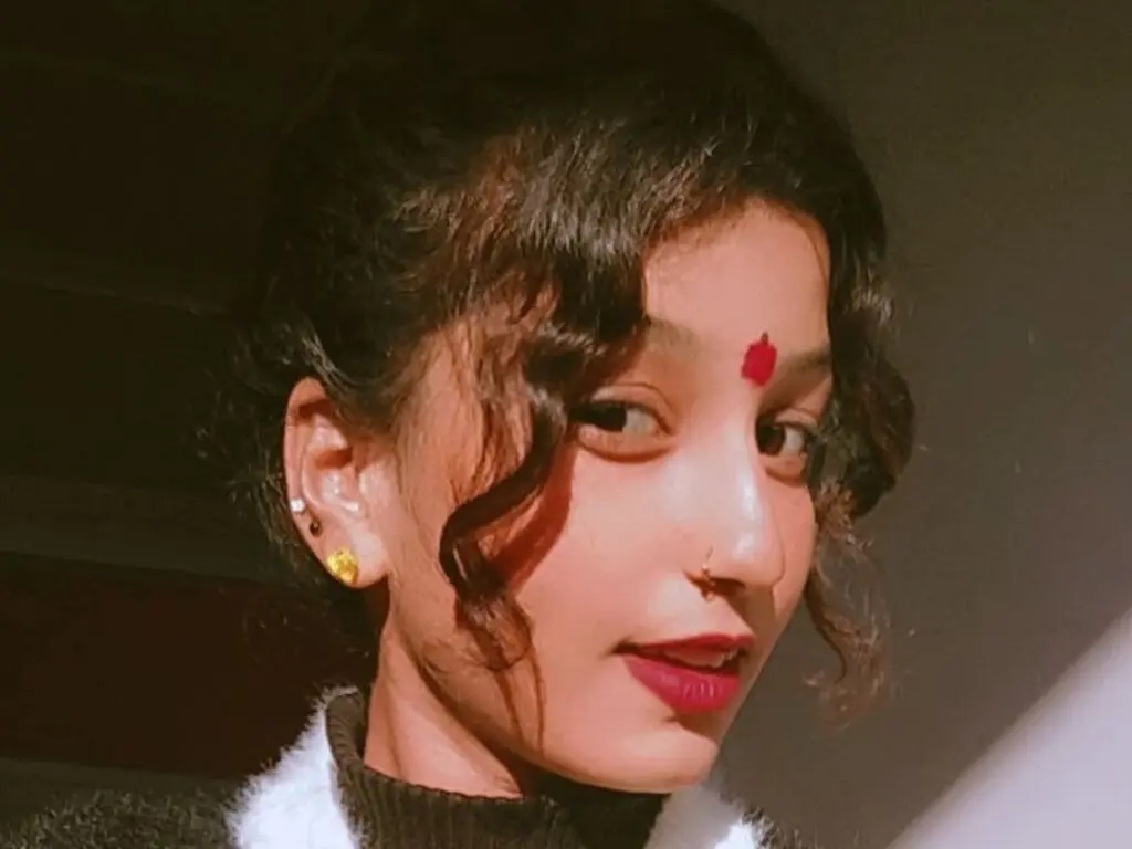 Young woman with curly hair and red bindi wearing a black sweater in warm indoor lighting.
