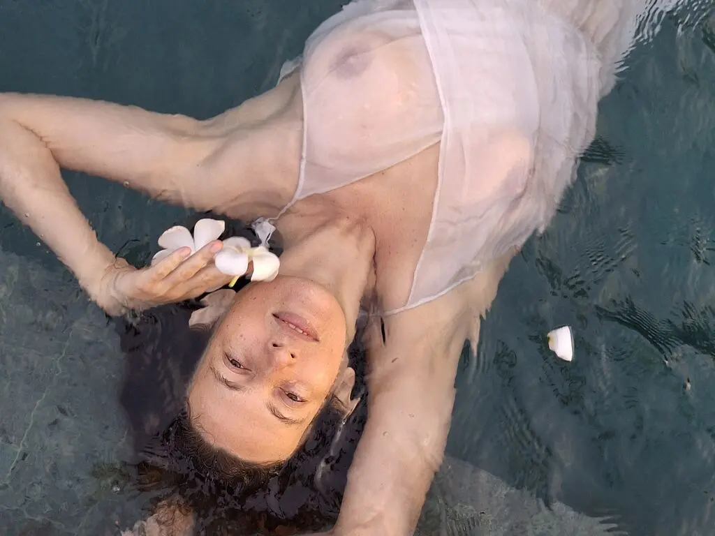 Serene woman in a white sheer dress floating peacefully in clear turquoise water with white flowers.
