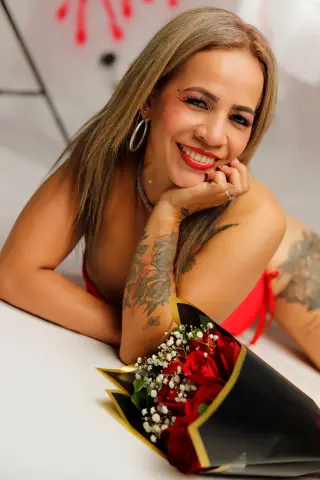 Smiling blonde woman in a red outfit lying next to a bouquet of red roses in a bright, romantic setting.